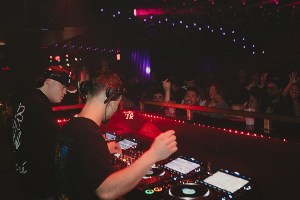 Jeremias and Arias behind the CDJs, lit red, playing to a packed room.