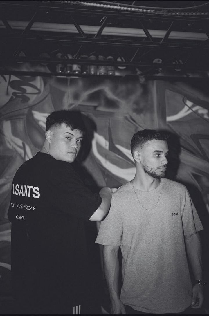 Black and white portrait of the duo standing in front of a graffiti wall under stage truss.